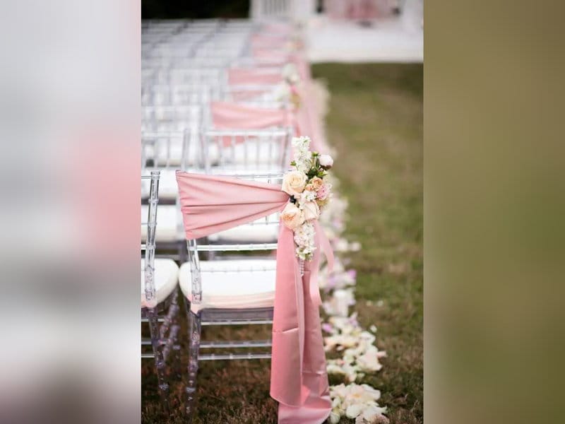 Rows of clear Chiavari chairs with pink sashes along a flower-lined aisle